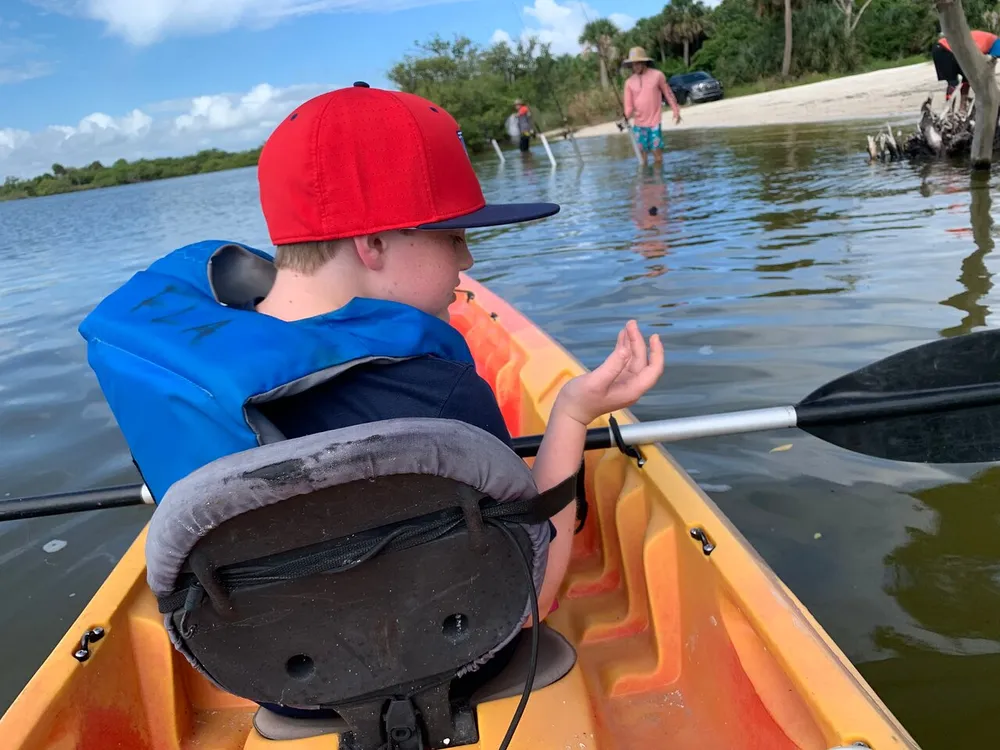 A child wearing a life jacket sits in a kayak gesturing with one hand while holding a paddle with people wading in the background near a sandy area