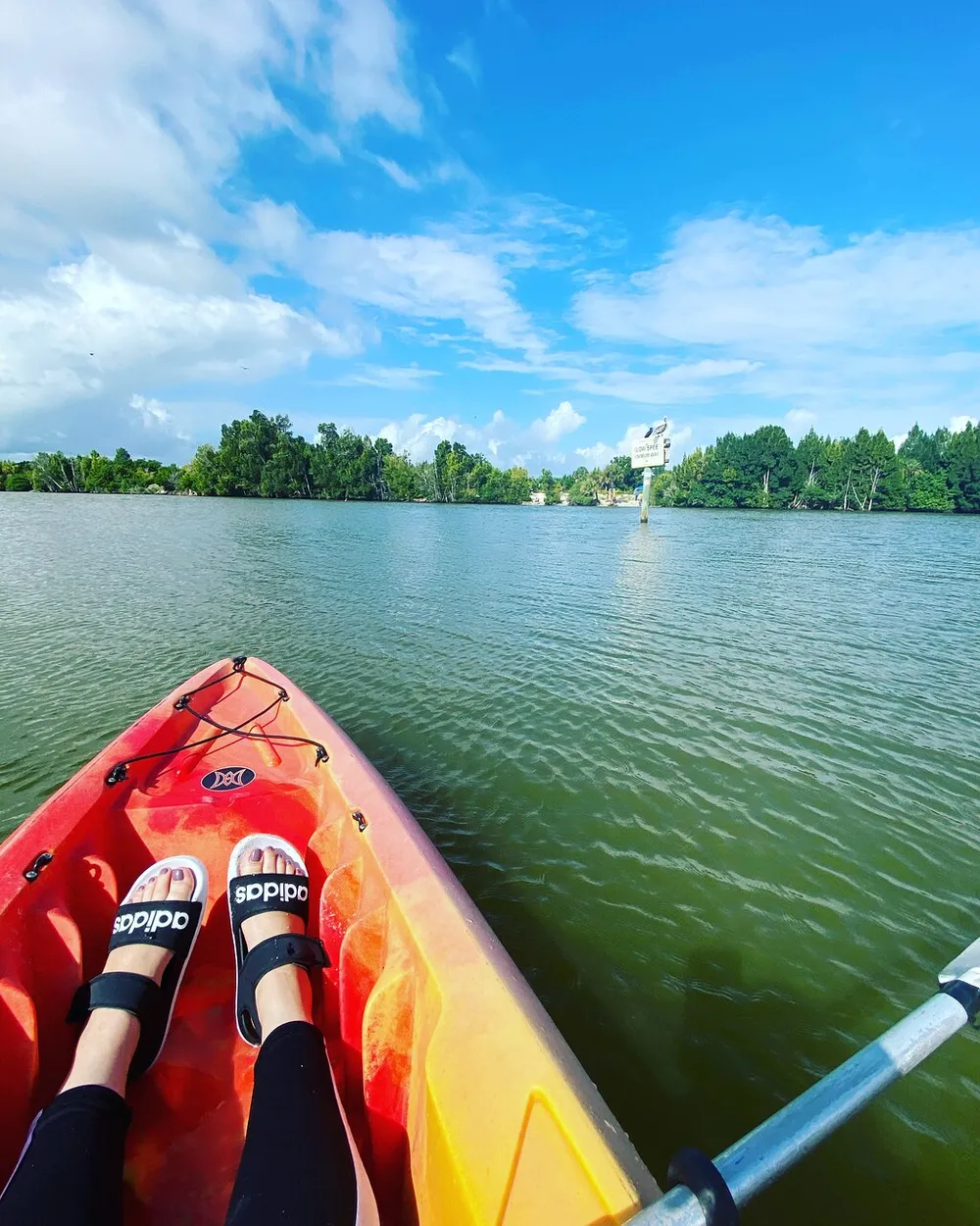 The image captures a first-person view of a person kayaking on a calm river with greenery on the banks and a mix of clouds and blue sky overhead