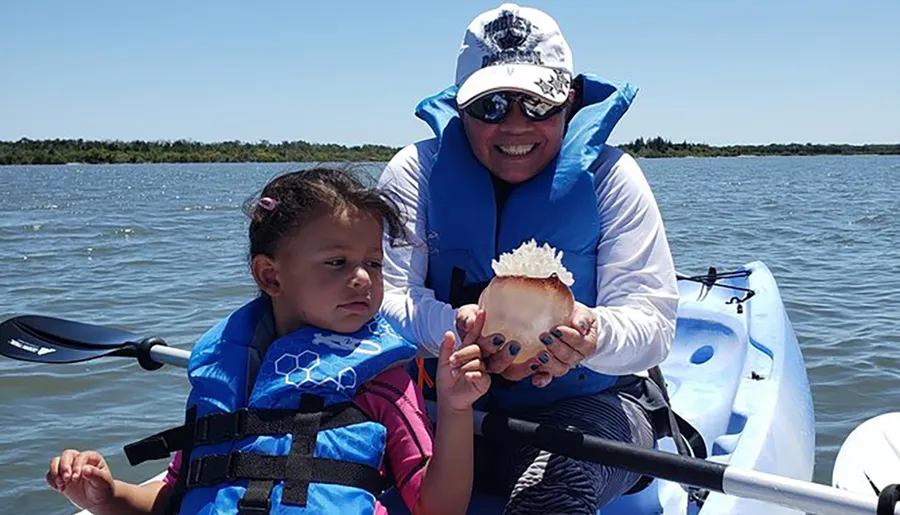 A child and an adult wearing life jackets are on a kayak, with the adult showing a sea creature to the child.