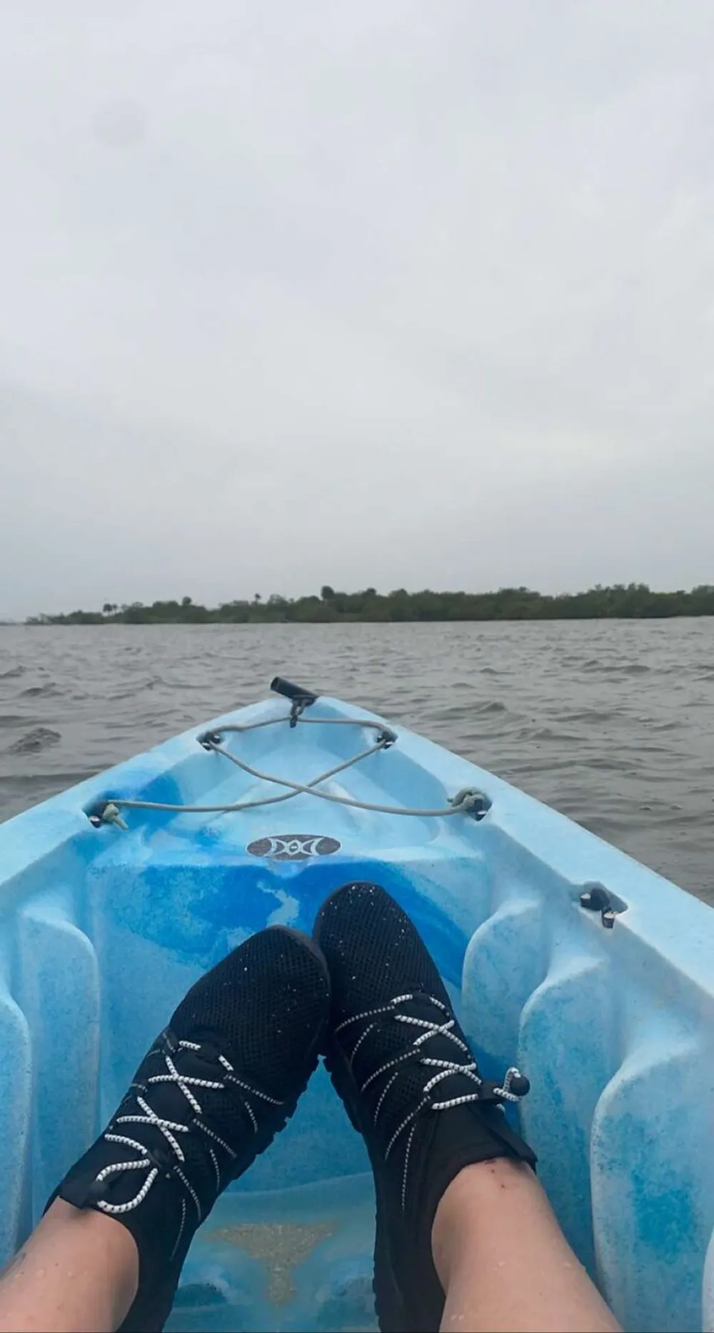 A person wearing water shoes is sitting in a blue kayak on a body of water with an overcast sky above