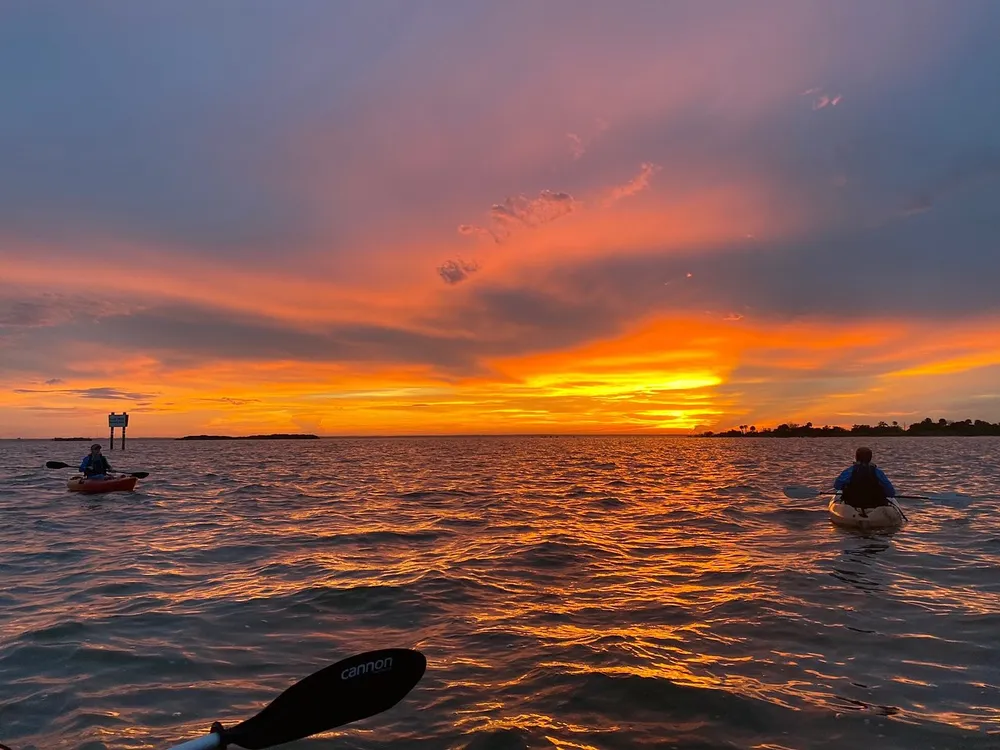 Two kayakers paddle on tranquil waters under a vibrant sunset sky