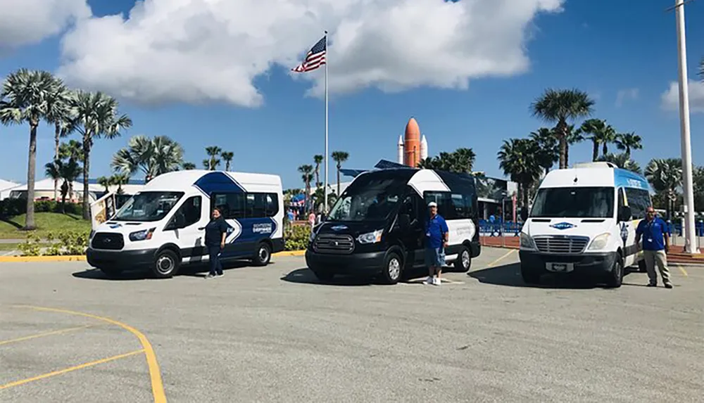Three shuttle buses are parked with drivers standing by them in a sunny parking lot with palms and a space shuttle display in the background under a clear blue sky