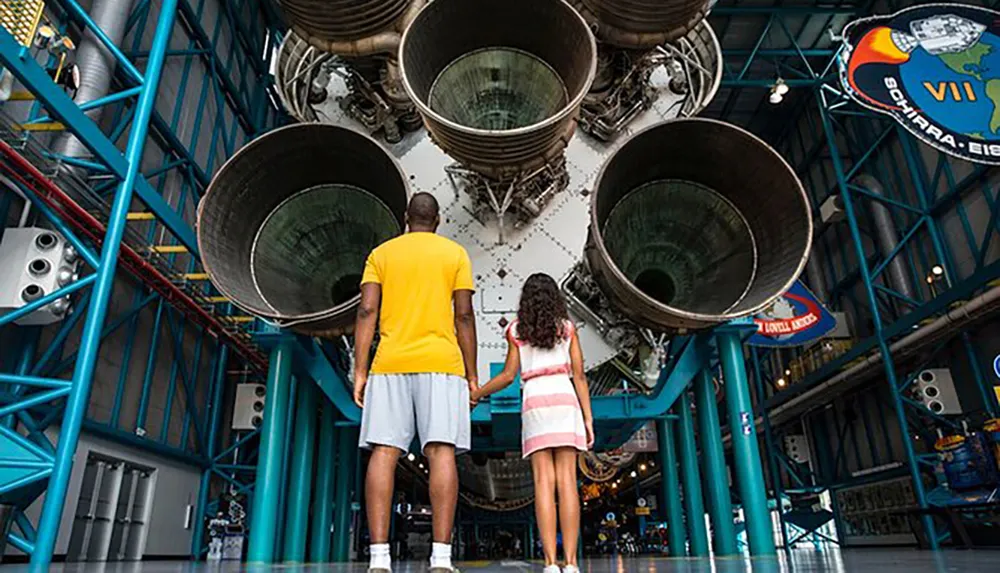 A man and a child are holding hands while standing in front of the massive engines of a Saturn V rocket displayed at a museum