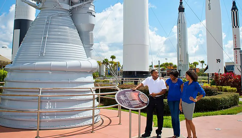 Three people are conversing near a large rocket engine exhibit with several rockets displayed in the background under a sunny sky
