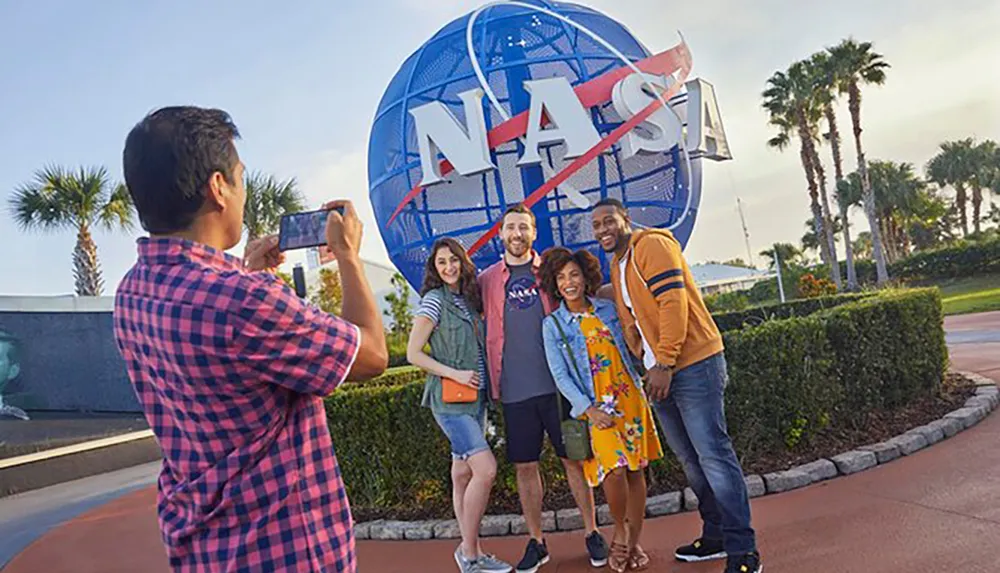 A group of friends is posing for a photo in front of a large NASA logo sign while another individual takes their picture