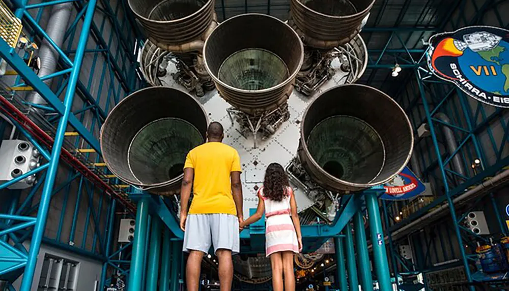 An adult and a child are standing hand in hand gazing up at the massive engines of a Saturn V rocket displayed at a space museum