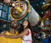 A smiling man and a young girl look up in awe at a spacecraft exhibit in a space museum
