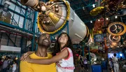 A smiling man and a young girl look up in awe at a spacecraft exhibit in a space museum.