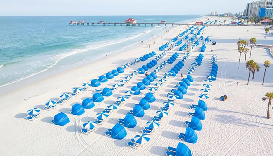 Aerial view of a sandy beach with neatly aligned rows of blue beach umbrellas and loungers, with people enjoying the seaside near a pier.