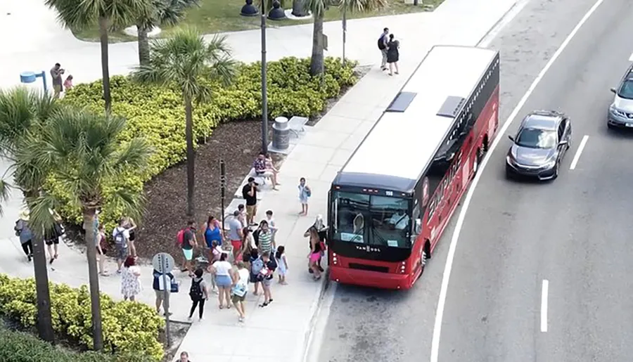 A group of passengers is boarding a red city bus at a stop, with cars passing by on the adjacent lane.