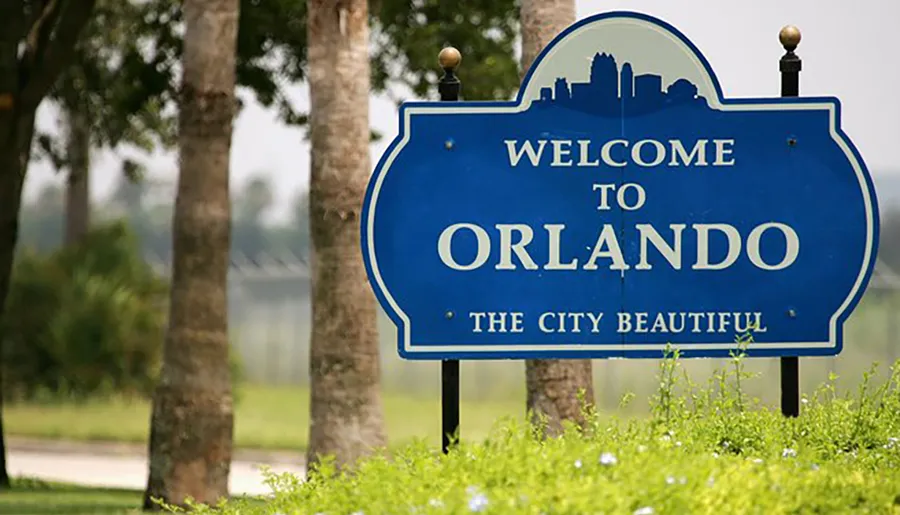 The image shows a blue welcome sign with white text reading Welcome to Orlando THE CITY BEAUTIFUL, surrounded by greenery and palm trees.