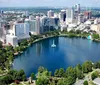 The image depicts a scenic view of a modern city skyline reflected in a tranquil body of water framed by lush greenery and a clear blue sky