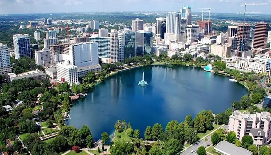 This image is an aerial view of a downtown skyline with modern buildings surrounding a serene lake which features a fountain in its center.