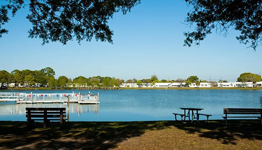 The image shows a serene lakeside scene with a dock, pedal boats, a wooden bench, a picnic table under trees, and a clear blue sky.