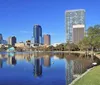 The image depicts a scenic view of a modern city skyline reflected in a tranquil body of water framed by lush greenery and a clear blue sky