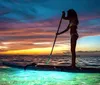 A silhouette of a person paddleboarding on a tranquil sea illuminated by underwater lights against a dramatic sunset sky