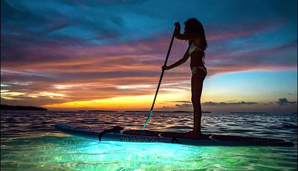 A silhouette of a person paddleboarding on a tranquil sea illuminated by underwater lights against a dramatic sunset sky