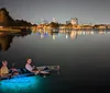 A silhouette of a person paddleboarding on a tranquil sea illuminated by underwater lights against a dramatic sunset sky