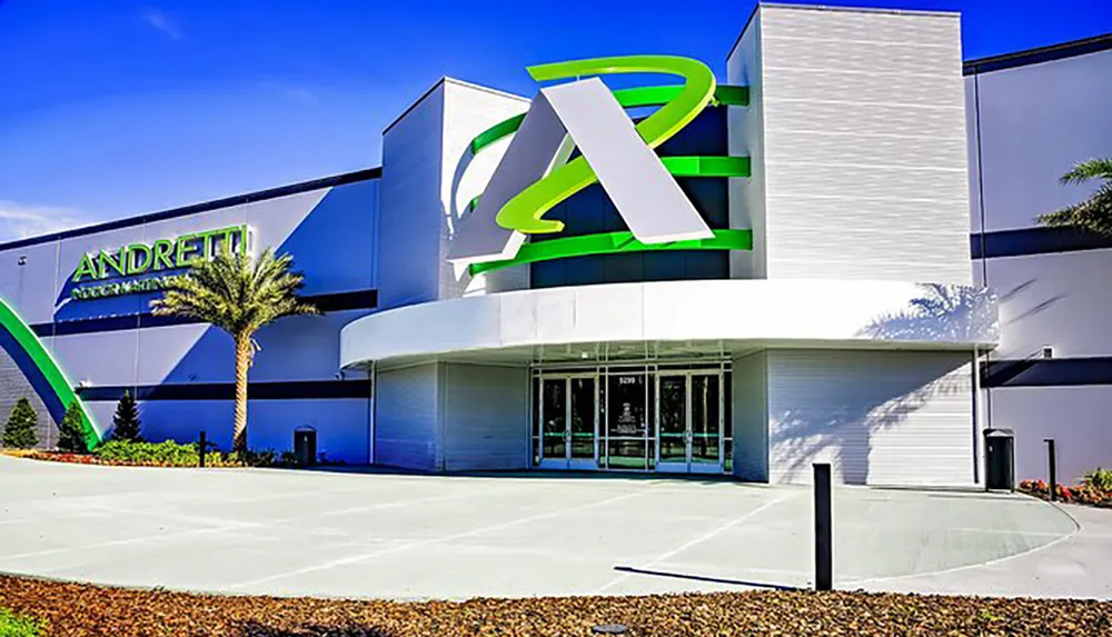 The image shows the modern facade of an Andretti Indoor Karting  Games entertainment complex featuring a large green and white logo under a blue sky