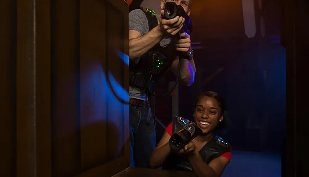 Two people are enjoying a game of laser tag equipped with vests and phasers in a dimly lit arena