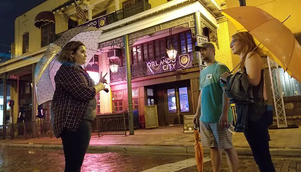 Four individuals stand on a city street at night two of them holding umbrellas with a building illuminated with purple lighting in the background