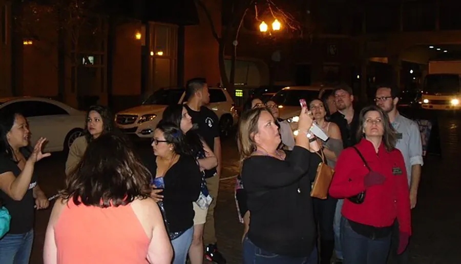 A group of people appear engaged in a nighttime outdoor activity, some looking attentive and others taking photos with their smartphones.
