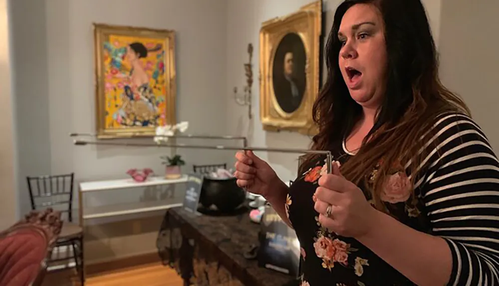 A surprised woman with an open mouth is holding a broken piece of artwork in a gallery with other pieces on display in the background