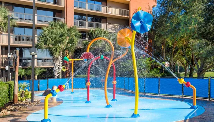 The image shows a colorful splash pad with water features in front of a multi-story resort building lined with palm trees under a clear blue sky