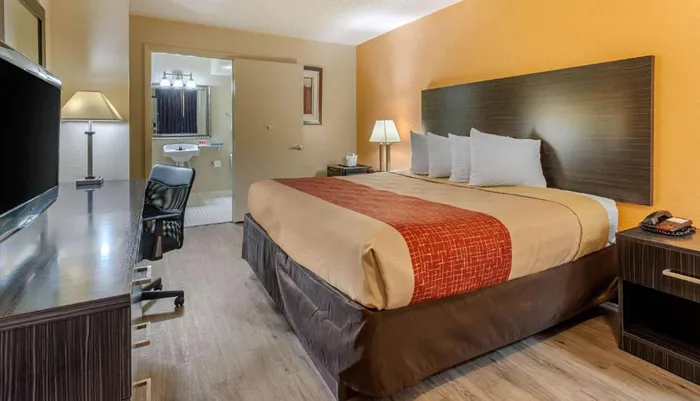 The image shows a neatly arranged hotel room with a large bed a desk with a chair a flat-screen TV and an open bathroom door in the background