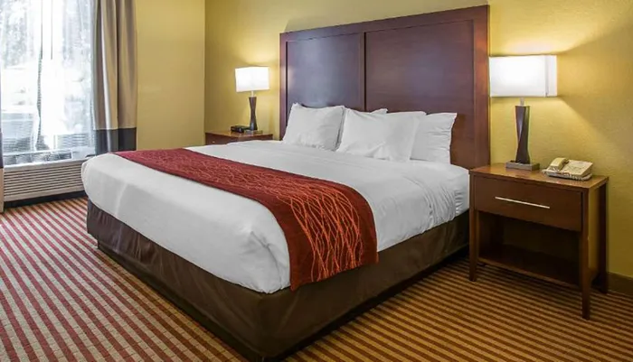 A neatly made king-size bed with white linens and a red throw blanket is the centerpiece of a warmly lit hotel room with striped carpeting and a yellow wall