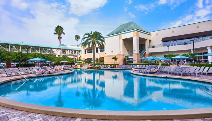 The image shows a serene resort swimming pool surrounded by lounging guests palm trees and a hotel structure in the background under a partly cloudy sky