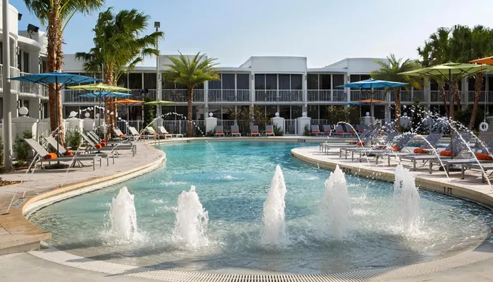 The image shows a tranquil hotel pool area with fountains surrounded by palm trees and loungers under a clear sky
