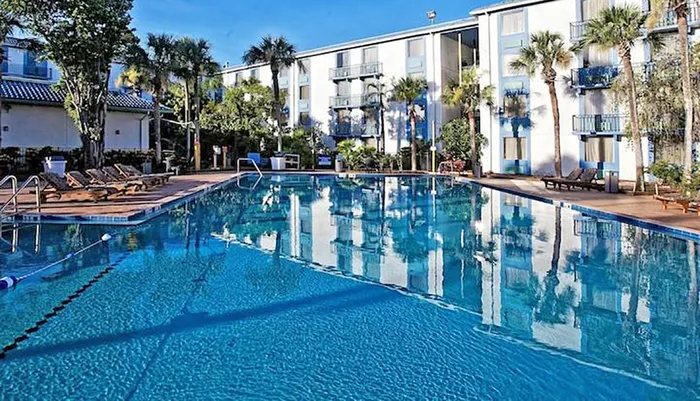 The image shows a calm and inviting outdoor swimming pool surrounded by loungers trees and a multi-story hotel building under a clear blue sky
