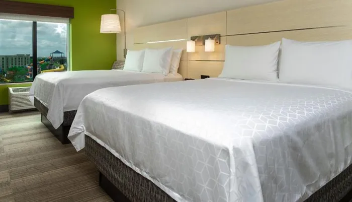 The image shows a bright and neatly arranged hotel room with two large beds a green accent wall modern lighting and a view of the outside from a large window