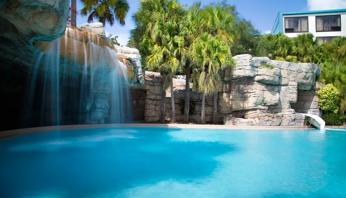 The image shows a serene swimming pool with a cascading artificial waterfall surrounded by tropical vegetation and architectural structures conveying a resort-like atmosphere
