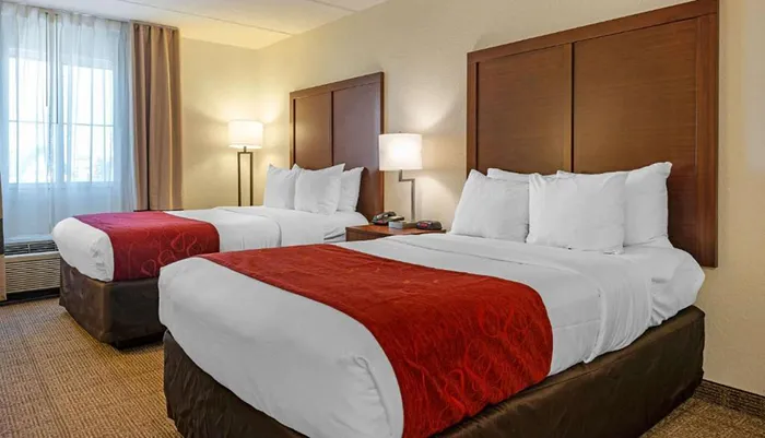 The image shows a neatly arranged hotel room with two double beds featuring white linens and red decorative bed runners flanked by nightstands and lamps with a window in the background