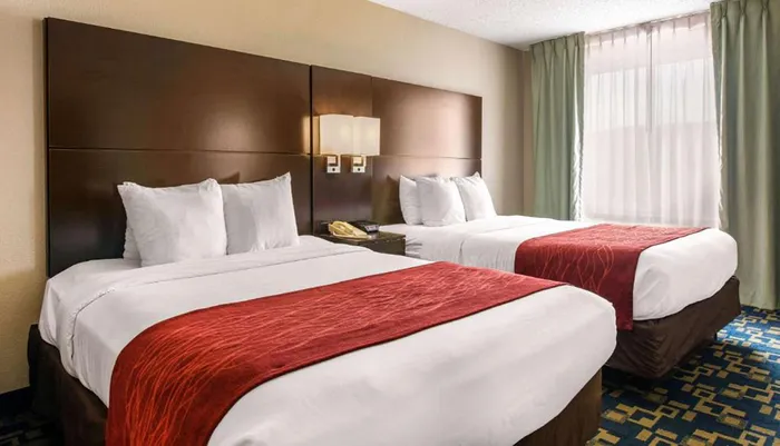 The image shows a tidy hotel room with two beds featuring white linens and red bed runners a headboard bedside lamps and a curtained window