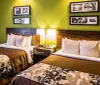 The image shows a double hotel room with two beds each with brown covers and floral-patterned throws against a lime green wall with dark wooden headboards a nightstand with a lamp and open doors to a bathroom and entryway in the background
