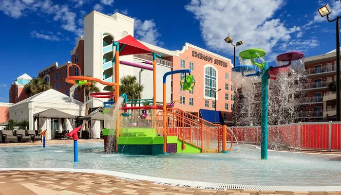 This image shows a vibrant and colorful childrens water play area with slides and splash buckets in front of a hotel building under a clear blue sky