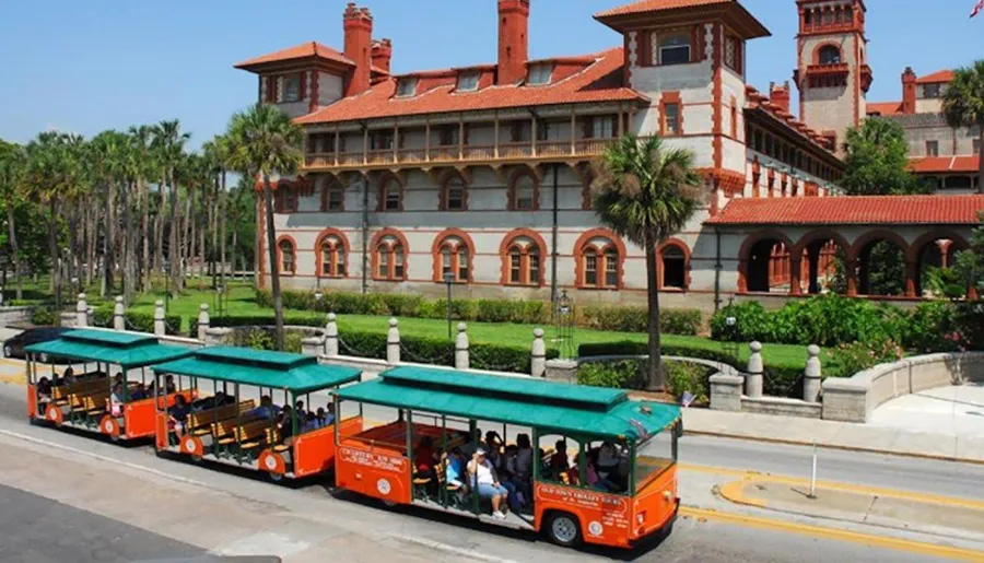 Orange trolley cars filled with tourists are passing by a historical red-roofed building surrounded by palm trees on a sunny day.