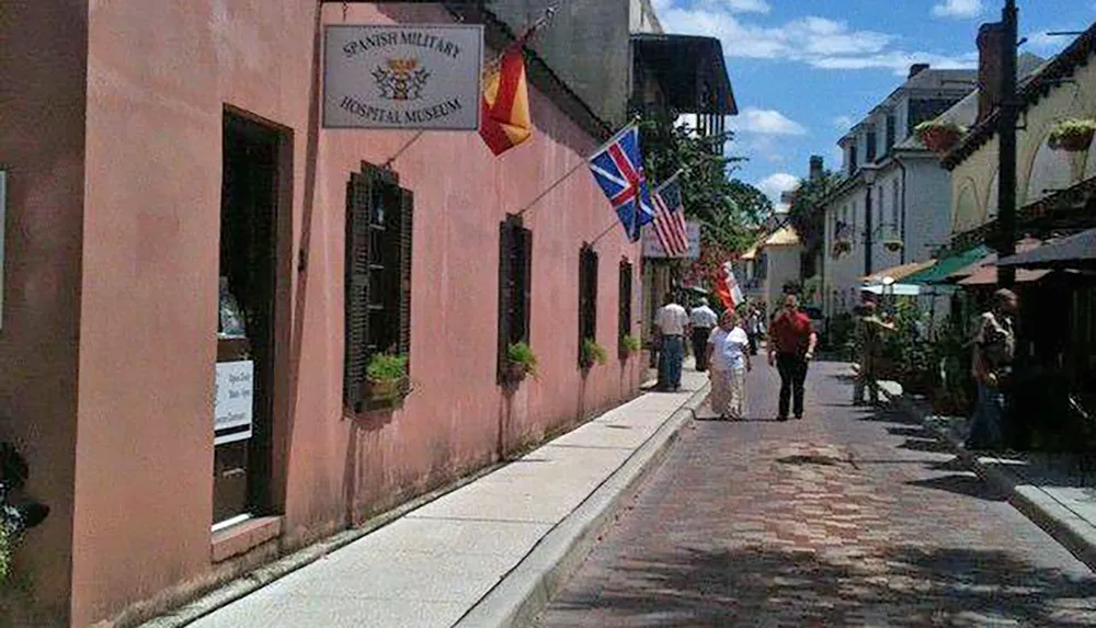 Pedestrians stroll past the pink facade of the Spanish Military Hospital Museum under a sunny sky flanked by various flags and complemented by the quaint architecture of a historic street