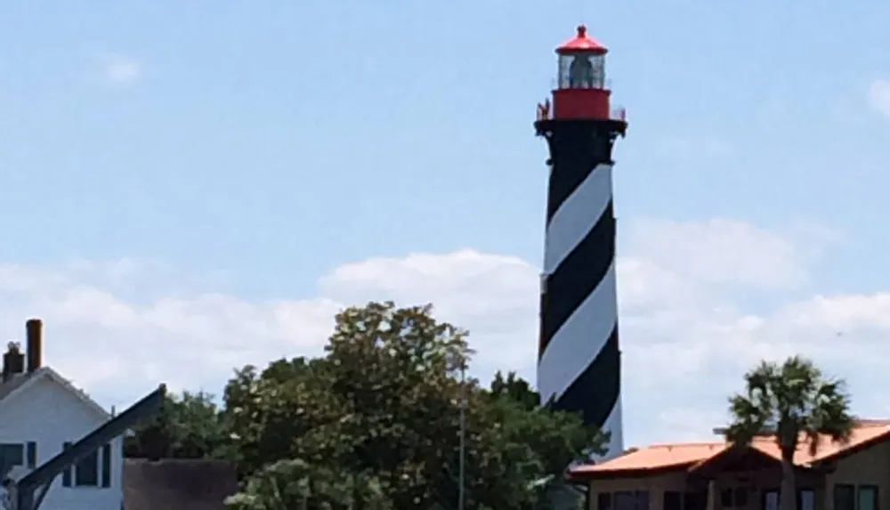 A tall spirally-striped black and white lighthouse with a red top stands prominently under a blue sky with scattered clouds with houses and trees in the foreground