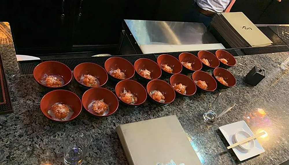 The image shows a series of small red bowls lined up on a bar counter each containing a similar serving of food with chopsticks resting on a holder and an empty glass nearby