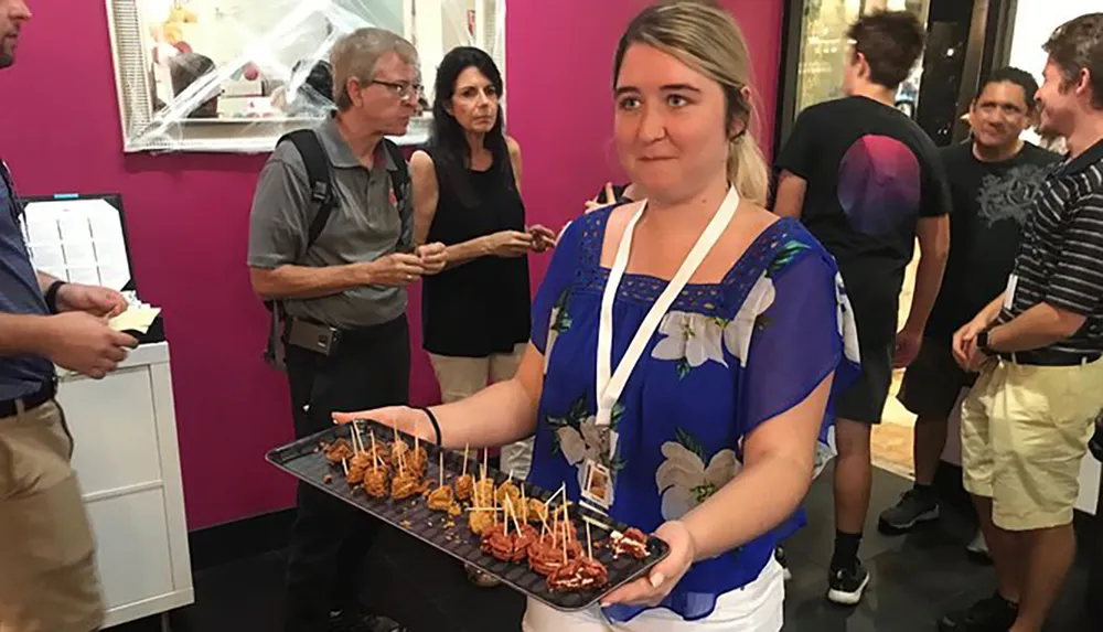 A woman holding a tray of appetizers looks at the camera with an amused expression as people behind her appear to be in a conversation or waiting to be served