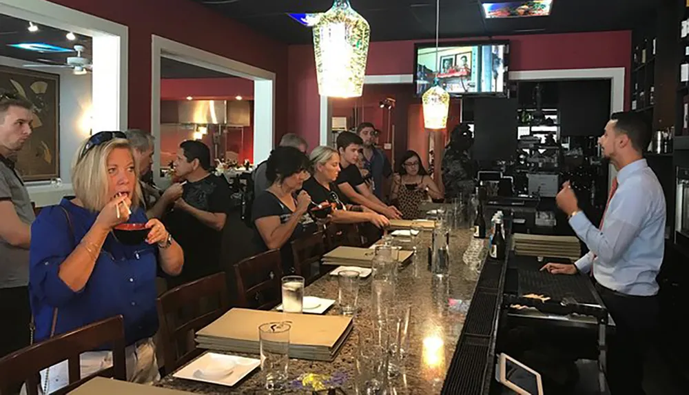 People are attentively watching a man speaking at the end of a bar in a restaurant while one person is having a drink