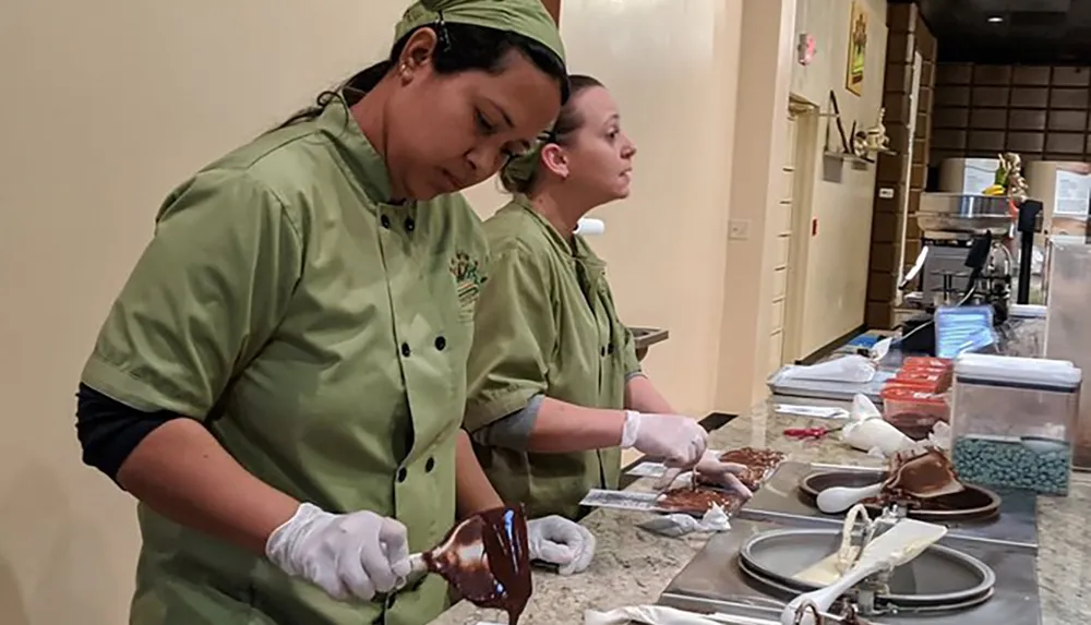 Two people in chef uniforms are working diligently in a kitchen one spreading chocolate on a dessert