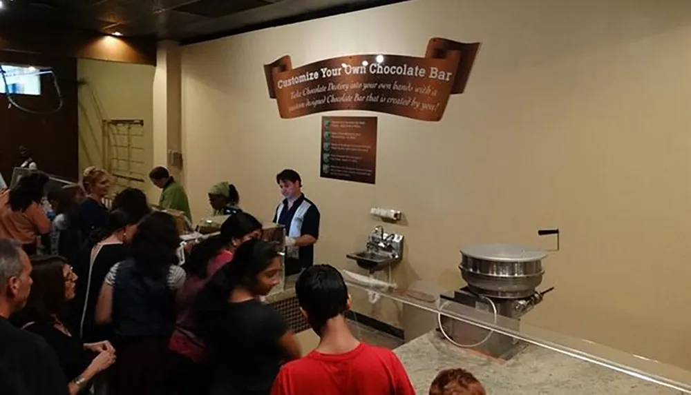 People are queued at a chocolate workshop where you can customize your own chocolate bar as indicated by the sign above