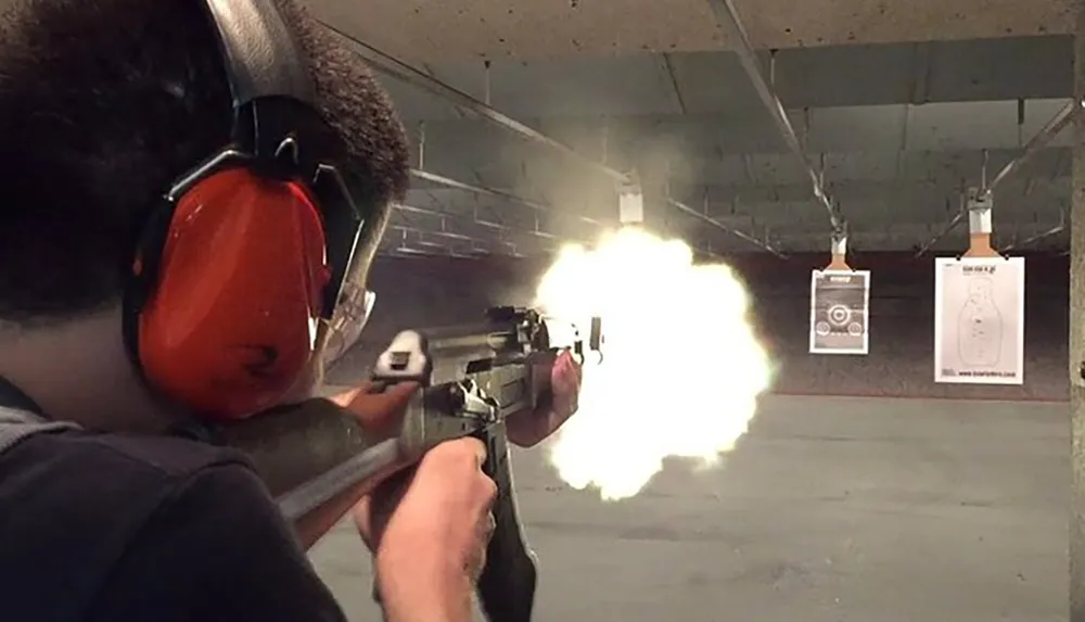 A person with ear protection is firing a rifle at an indoor shooting range with a visible muzzle flash coming from the barrel