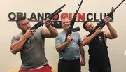 Three men are posing with firearms in front of a wall that has 