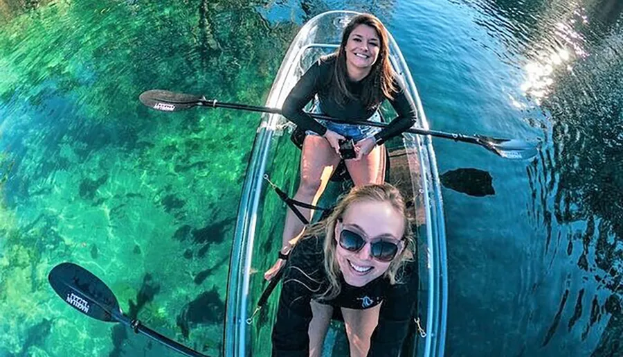 Two women are smiling and enjoying a clear water kayaking experience in a transparent kayak.